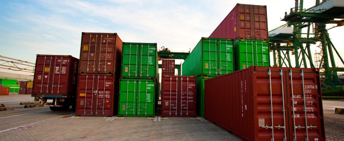 Contenedores de carga apilados en un puerto marítimo, en tonos rojos y verdes, con grúas industriales al fondo y cielo parcialmente despejado