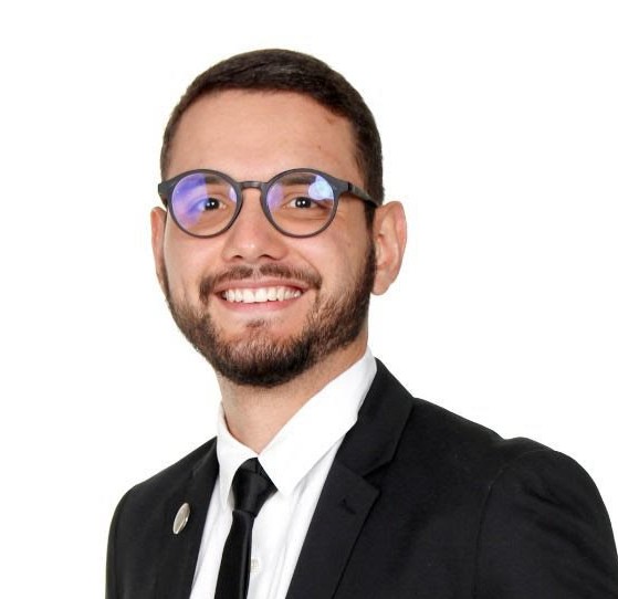 Retrato profesional de un hombre joven con barba corta y gafas redondas, vestido con traje formal negro, camisa blanca y corbata negra. Sonríe y mira a la cámara sobre un fondo blanco, proyectando una imagen profesional y accesible