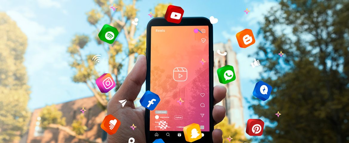 Primer plano de una mano sosteniendo un teléfono móvil que muestra la pantalla de creación de Reels en una red social. Alrededor del dispositivo flotan íconos tridimensionales de varias plataformas digitales como Instagram, Facebook, YouTube, WhatsApp, Pinterest, Messenger, entre otros. El fondo muestra árboles y edificios desenfocados bajo un cielo azul, simbolizando la conectividad digital y el auge de las redes sociales
