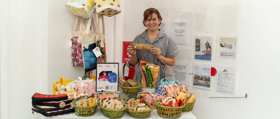 Mujer sonriente de pie detrás de una mesa que exhibe diversos productos artesanales. Sobre la mesa hay cestas de palma verde que contienen alimentos decorativos en forma de pupusas, tamales y otros platillos típicos salvadoreños hechos en tela. También hay bolsos y estuches coloridos. Al fondo, en la pared, se observan más bolsos colgados y un panel informativo con afiches y documentos institucionales. La escena tiene lugar en un espacio interior con paredes blancas, en un contexto de exposición o feria de emprendimientos.