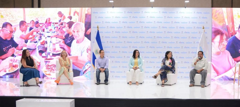 Panel de seis personas sentadas en un escenario durante un evento organizado por CONAMYPE y FANTEL. El fondo presenta una pantalla gigante con una imagen de una comida comunitaria, junto a un banner institucional con logotipos oficiales. Las banderas de El Salvador y CONAMYPE enmarcan el escenario, en un ambiente de diálogo y participación ciudadana.