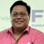 Hombre sonriente con camisa fucsia y gafas, posando frente a una pantalla con el logotipo parcialmente visible de una fundación. Luce un bolígrafo en el bolsillo y lleva una cadena dorada. La imagen transmite cercanía y profesionalismo.