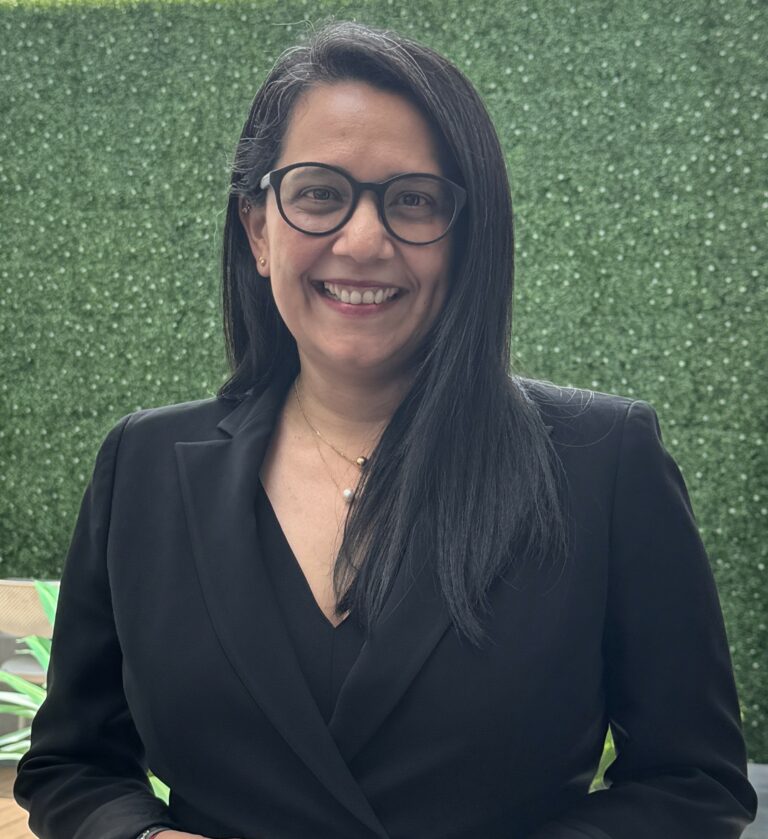Mujer sonriendo, vestida con traje formal negro y gafas, de pie frente a un fondo de vegetación artificial. Luce el cabello largo y lacio, y lleva un collar con detalles dorados. La imagen transmite profesionalismo y cercanía.