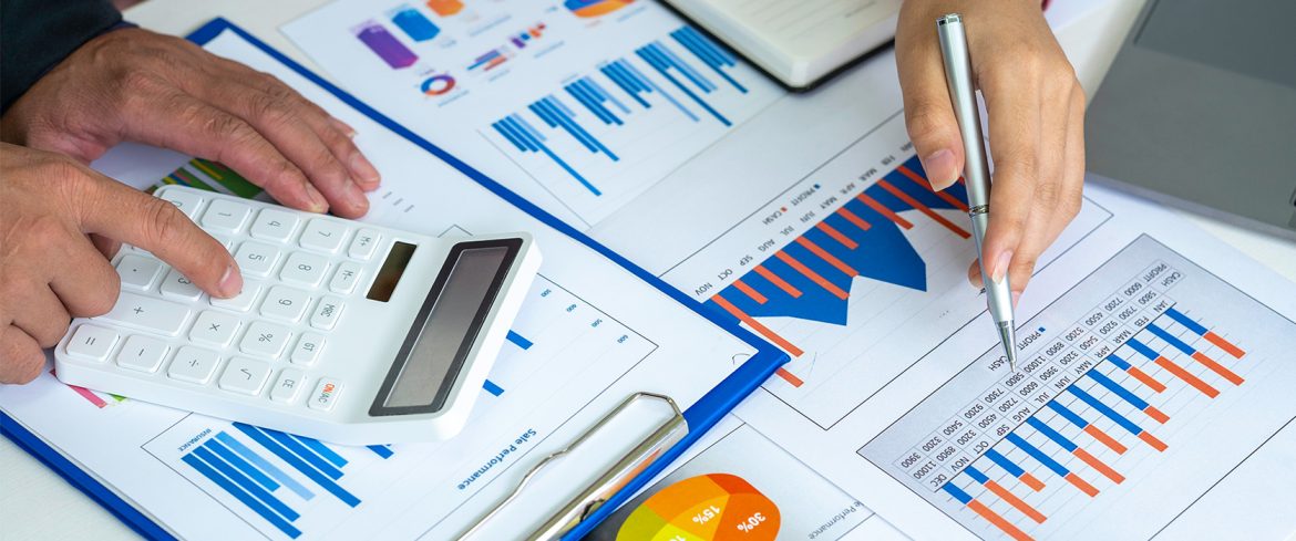 Personas analizando gráficos financieros y de rendimiento empresarial con una calculadora y bolígrafo sobre documentos que muestran barras comparativas y gráficos circulares. La imagen representa una revisión detallada del flujo de caja y la toma de decisiones financieras.