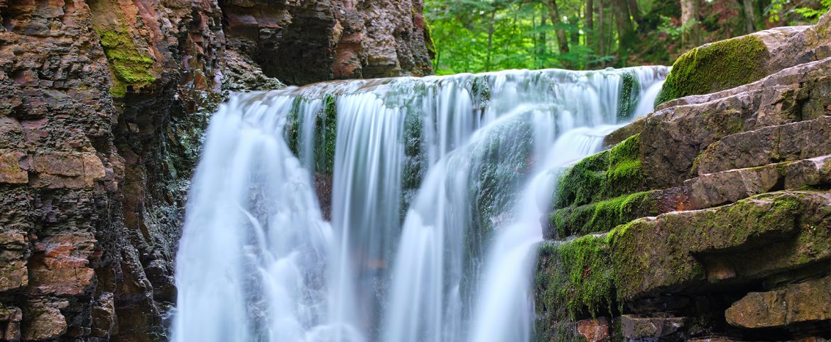 Cascada de agua cristalina descendiendo entre formaciones rocosas cubiertas de musgo, rodeada de un frondoso bosque verde. La escena transmite tranquilidad y riqueza natural, representando un entorno turístico de belleza escénica.