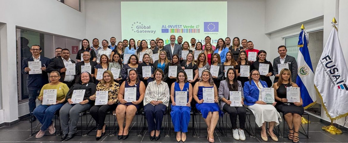 Fotografía grupal de graduación con más de 50 personas, entre hombres y mujeres, sosteniendo sus diplomas frente a una pantalla con los logotipos de Global Gateway, AL-INVEST Verde, Unión Europea y FUSAI. A la derecha, se observa la bandera de El Salvador y la bandera institucional de FUSAI. La imagen refleja un acto de cierre de formación con enfoque en sostenibilidad empresarial.