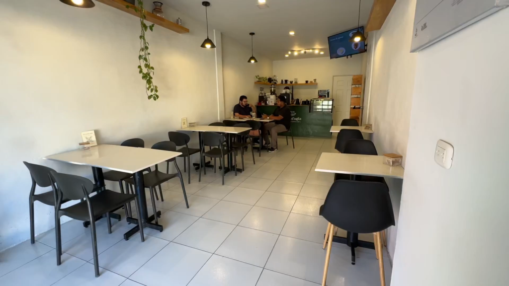 Interior del café “El Refugio” en Ahuachapán, con mesas y sillas negras organizadas en un espacio moderno y minimalista. Dos personas conversan en una mesa cercana a la barra, donde se observa el área de preparación de café con utensilios y estanterías. El lugar está bien iluminado, con lámparas colgantes, plantas decorativas y ambiente acogedor.