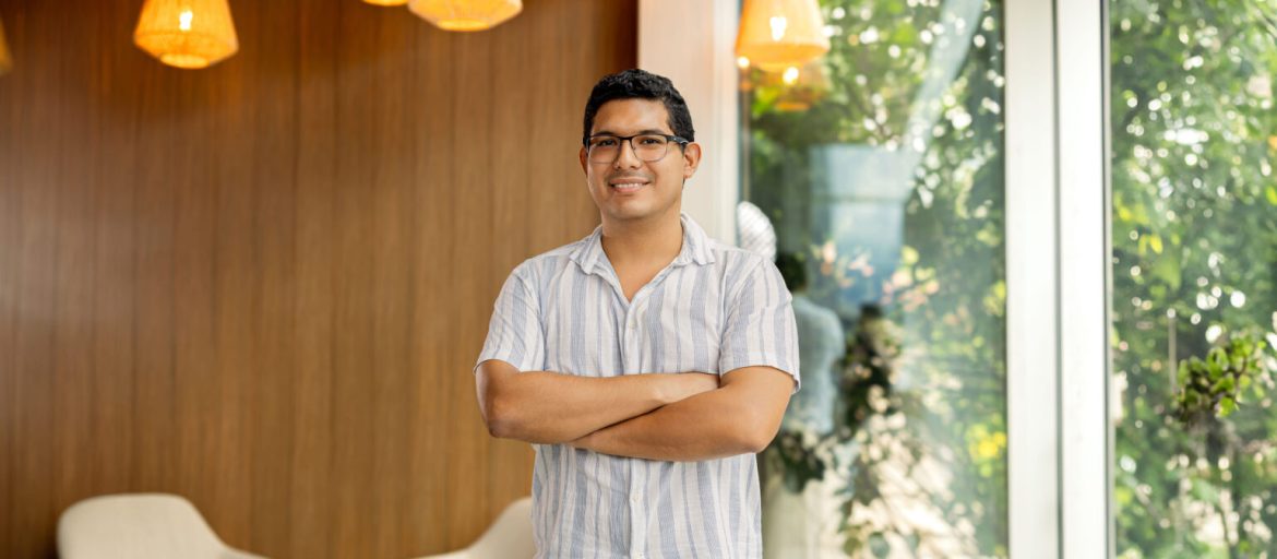 Hombre joven salvadoreño con gafas y camisa de rayas claras, posando con los brazos cruzados y una sonrisa en un espacio interior moderno con paredes de madera y luz natural que entra por una ventana con vista a árboles.