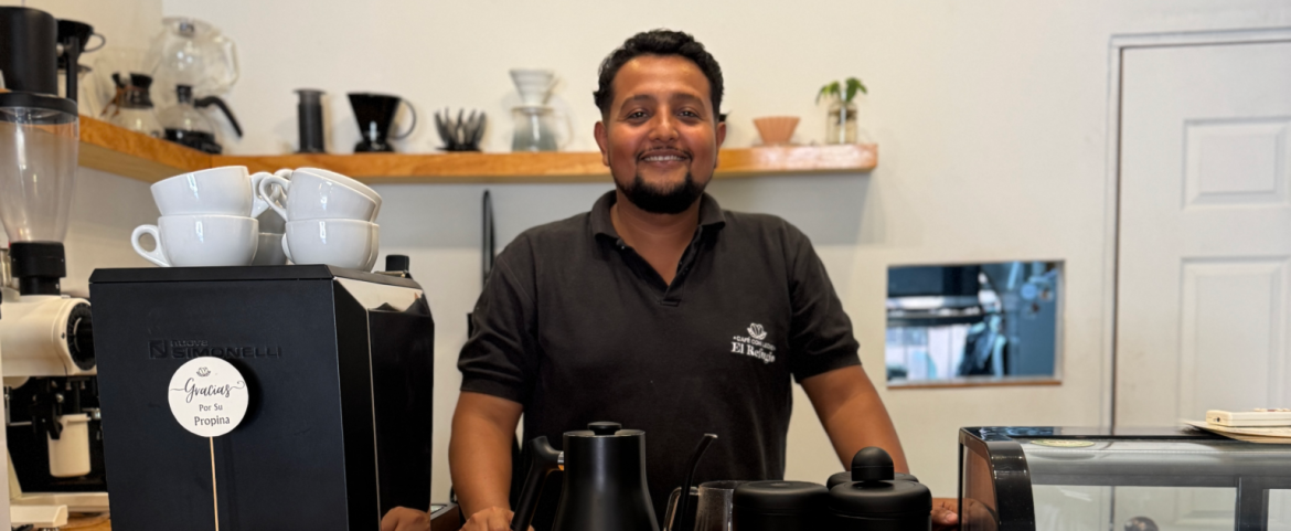 Hombre salvadoreño sonriente con barba corta y camiseta tipo polo con el logo de “Café Colaborativo El Refugio”, de pie detrás de la barra de una cafetería artesanal. Frente a él hay utensilios para preparar café y una máquina con un cartel que dice “Gracias por su propina”. Al fondo se observan tazas, cafeteras y equipos de barismo en un ambiente acogedor.