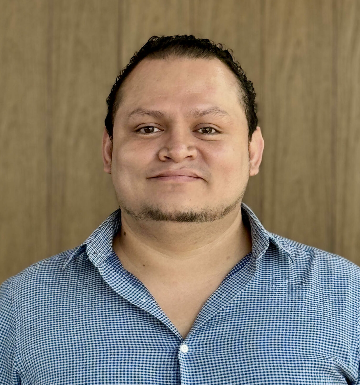 Hombre salvadoreño de rostro amable con camisa celeste de cuadros pequeños, posando frente a una pared de madera clara. Sonríe levemente a la cámara.