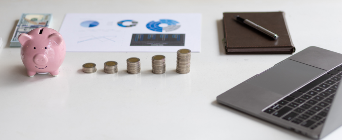 Alcancía rosa en forma de cerdito junto a cinco pilas crecientes de monedas, billetes, una hoja con gráficos financieros, una libreta con bolígrafo y una computadora portátil sobre un escritorio blanco. La imagen transmite conceptos de ahorro, planificación financiera y crecimiento económico.