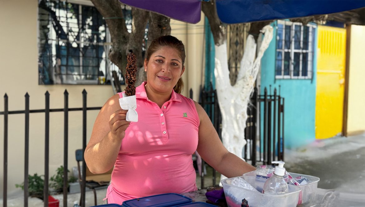 Mujer sonriente con blusa rosa sostiene un postre cubierto de chocolate en un palito, mientras atiende un puesto callejero con envases de hielo y desinfectante sobre la mesa.