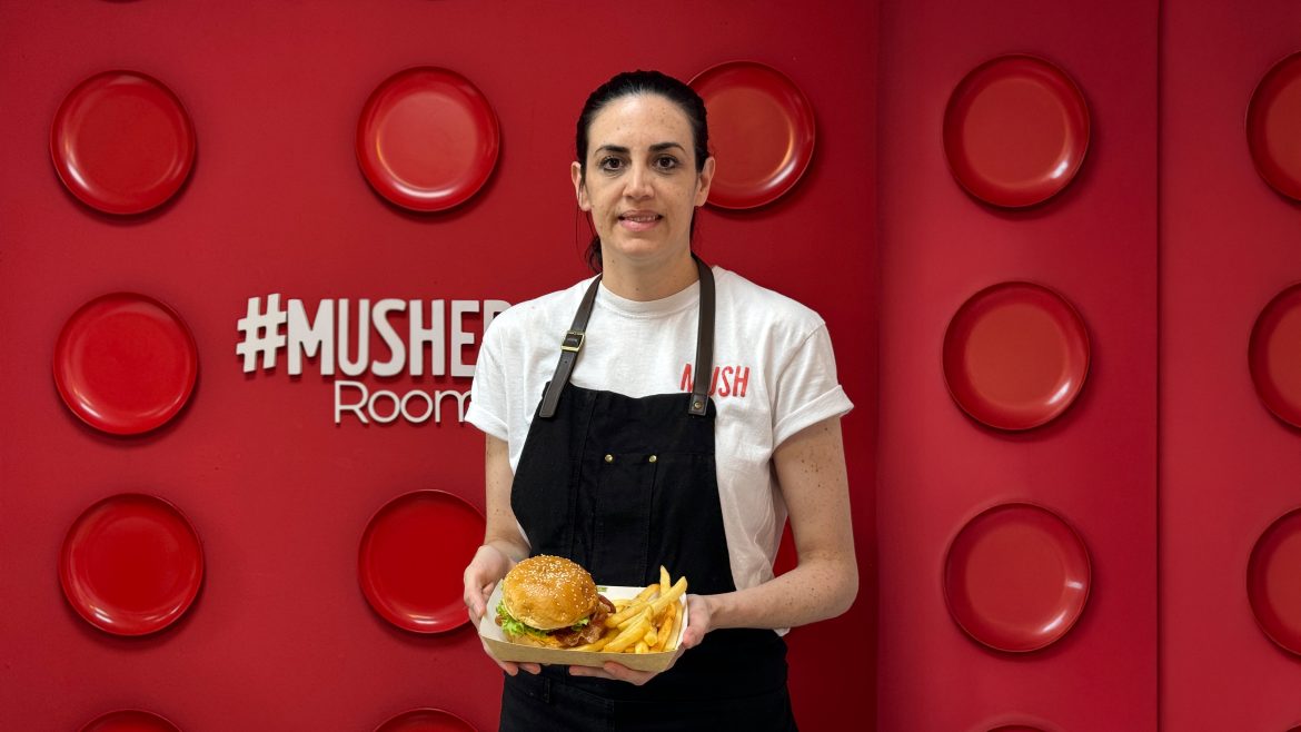 Mujer con camiseta blanca y mandil negro sostiene una hamburguesa con papas fritas, posando frente a una pared roja decorada con platos y el logo #MUSH