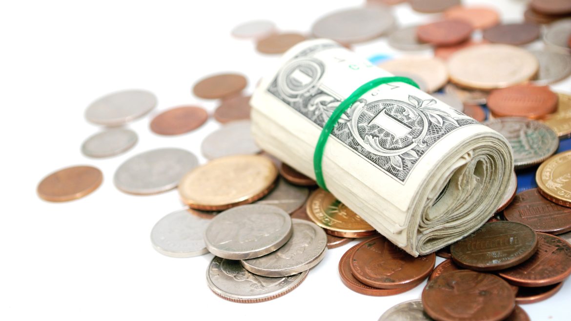 Billetes de dólar enrollados con una banda elástica verde sobre una superficie llena de monedas de diferentes denominaciones, representando ahorro, finanzas personales y economía.